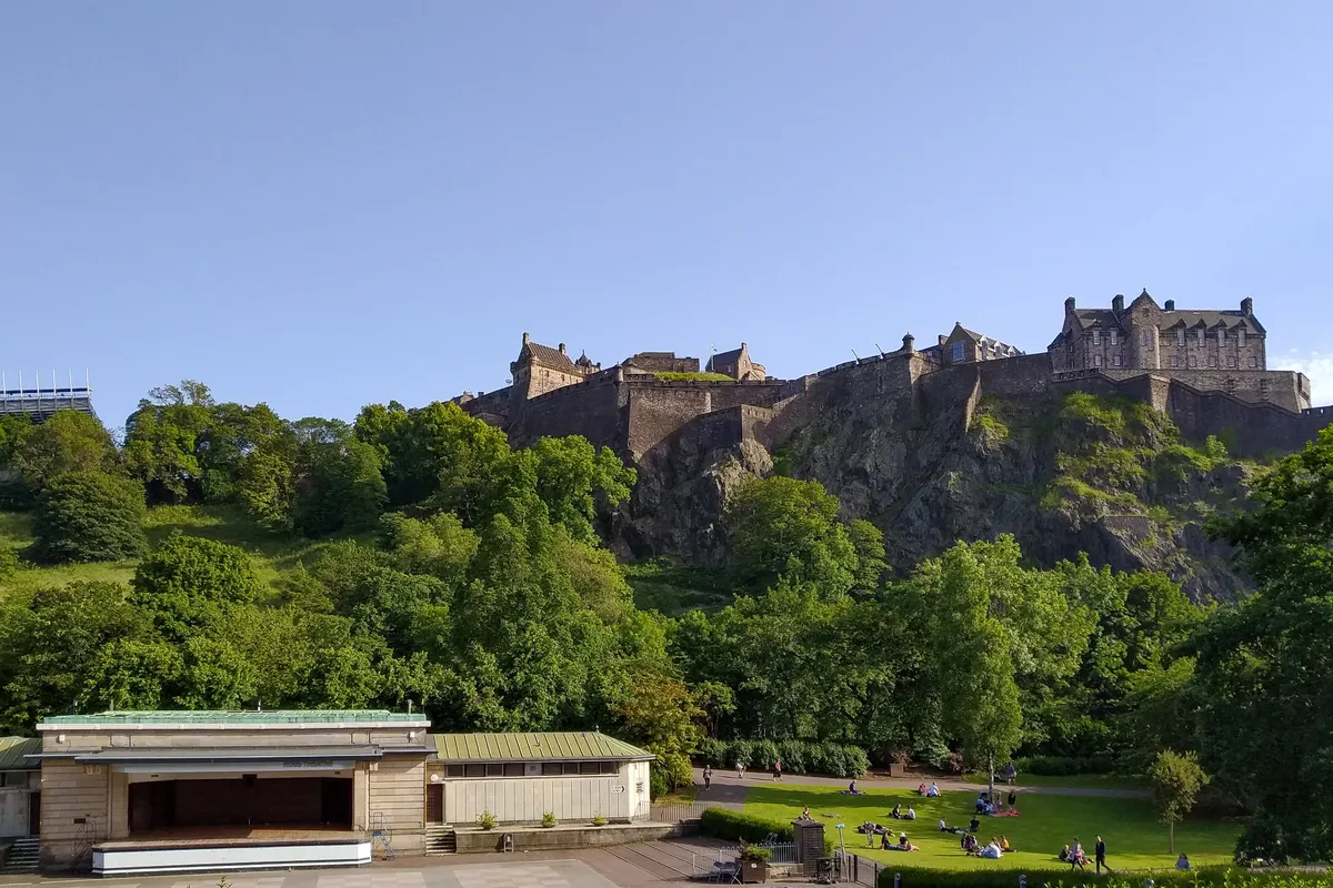 Castelo sobre um rochedo visto dos jardins, com torres, telhados, muralhas, vegetação e um coreto no primeiro plano.