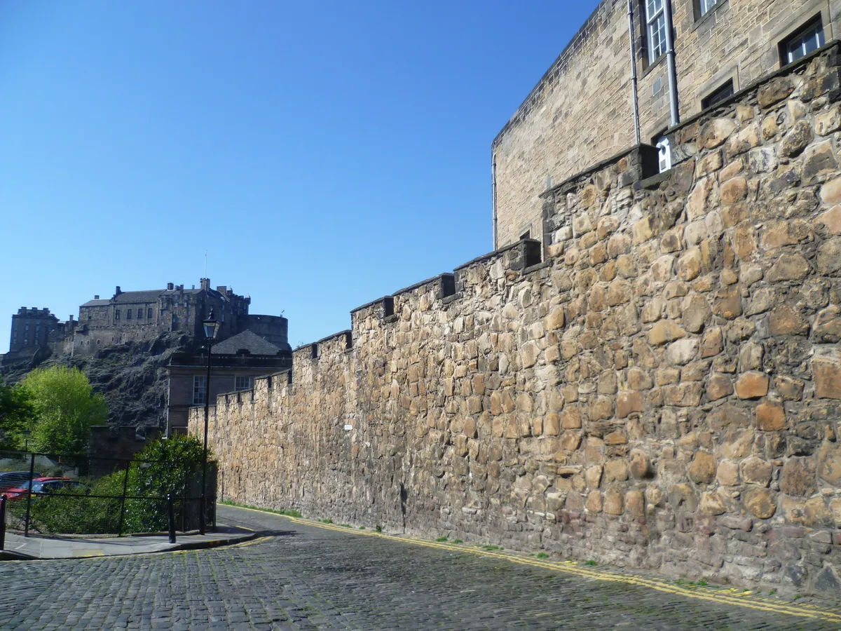 Seção de muralha de pedra junto de uma viela, com postes e vista para o castelo ao fundo.
