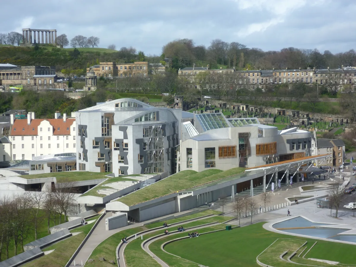 Vista ampla dos pavilhões do Parlamento Escocês, com telhados, janelas, espaços verdes e edifícios antigos ao fundo.