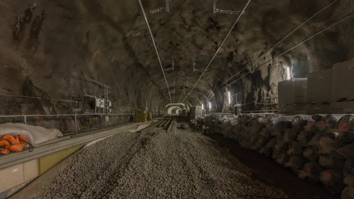 Túnel em obras com trilhos, lastro, cabos, tubos nas paredes e abertura iluminada ao fundo.
