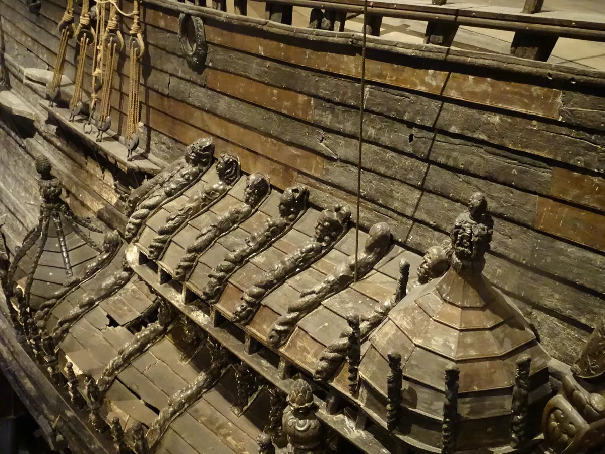 Detalhe do navio de madeira, com telhados inclinados, esculturas, cordas e tábuas no convés.