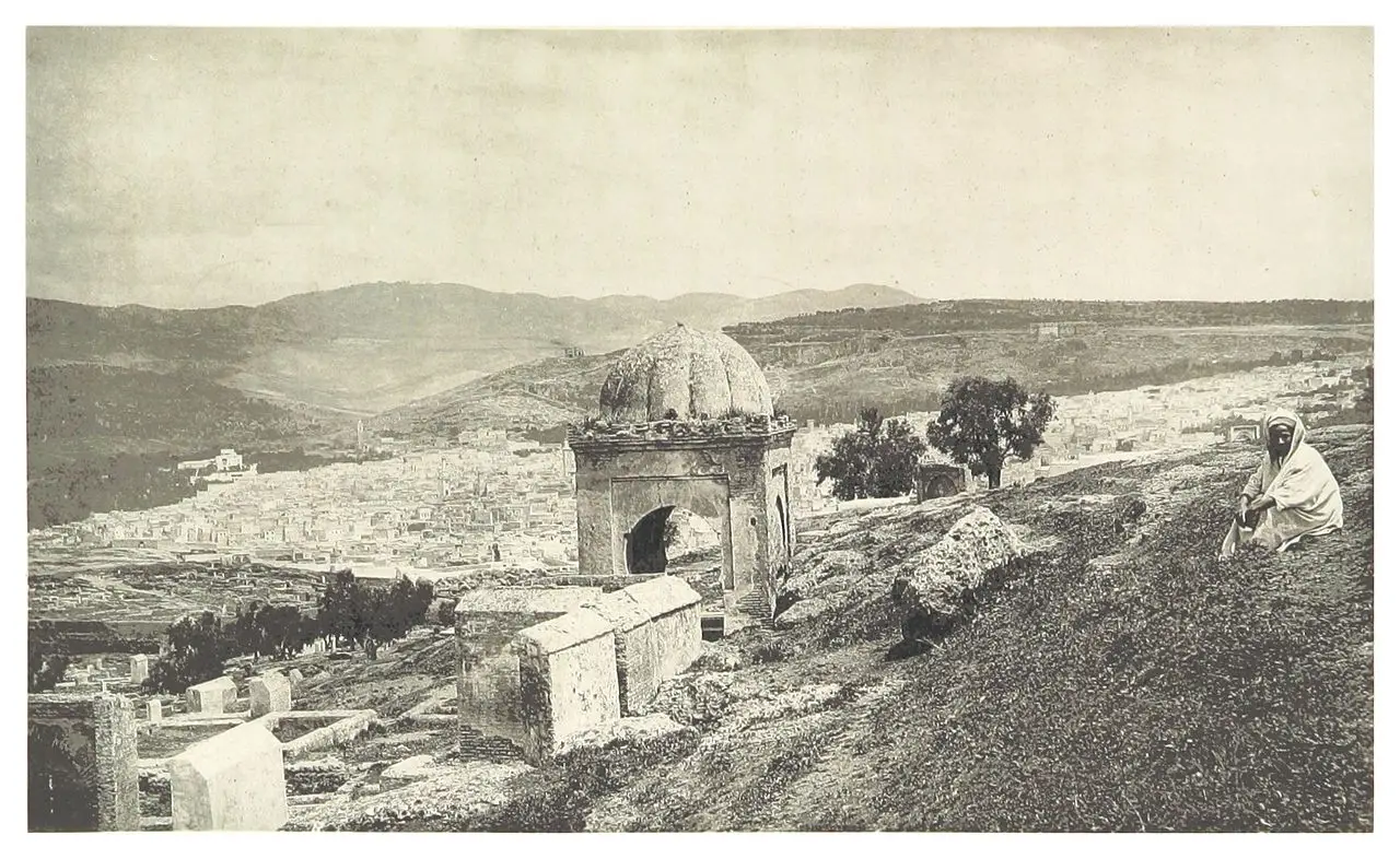 Fotografia em sépia de um mirante, com um pequeno edifício de cúpula em primeiro plano, um homem sentado à direita e a cidade clara num vale entre colinas.