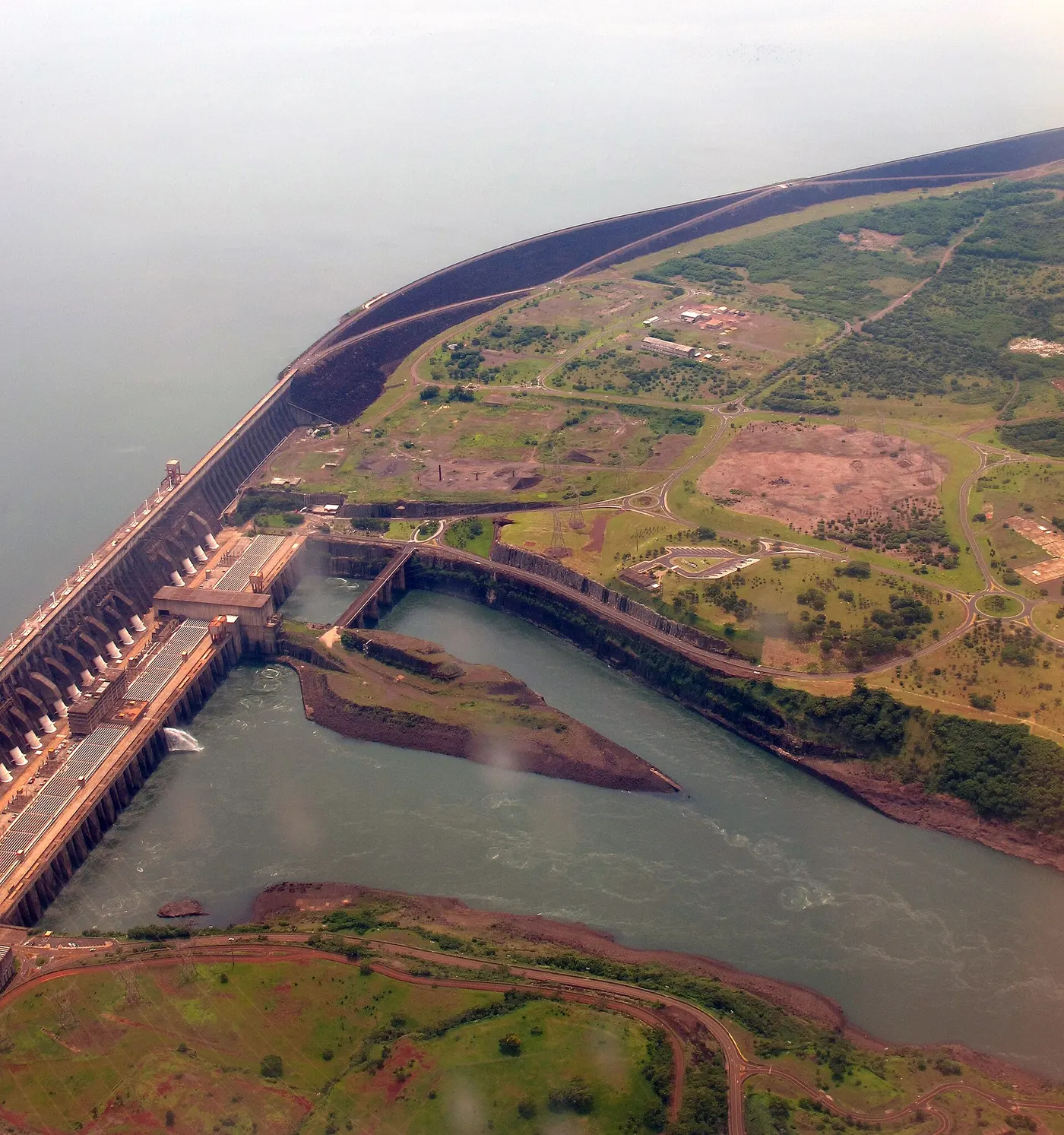 Vista aérea de uma barragem comprida, com água de um lado e o leito do rio do outro, sem pessoas.