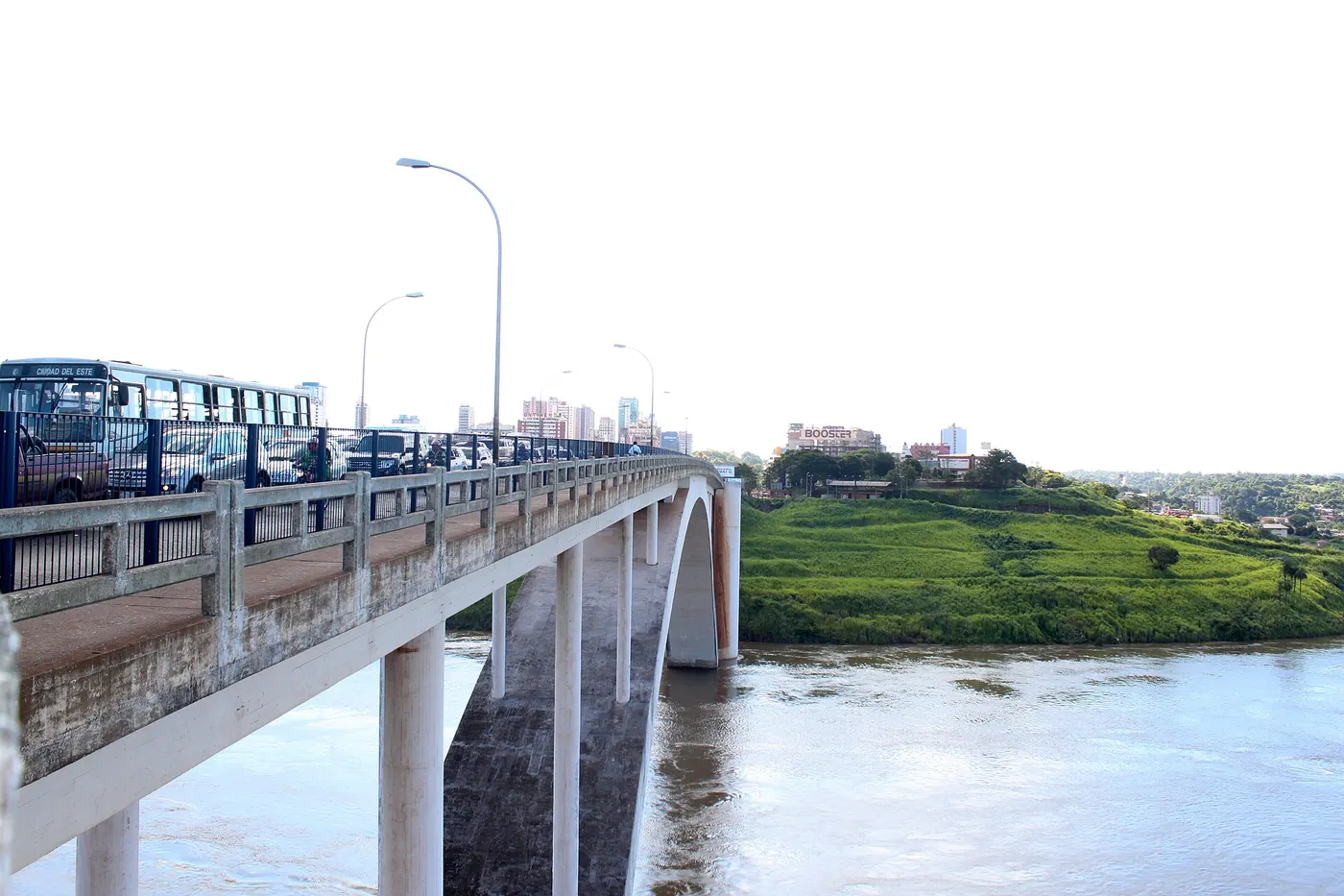 Uma ponte de arcos de concreto sobre um rio barrento, com ônibus e carros na pista.