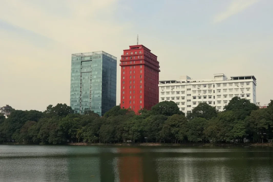 O lago Hoàn Kiếm, a faixa de árvores e as torres ao fundo, sob um céu claro.
