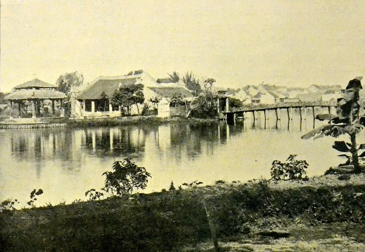 Fotografia em sépia do lago, com um pavilhão de telhado pontudo, casas baixas na margem e uma ponte de madeira sobre a água.