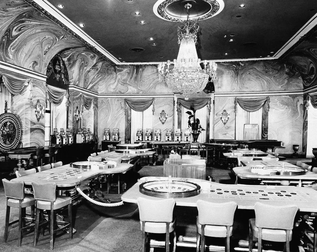 Uma sala de cassino vazia, com mesas de roleta, cadeiras e um lustre.