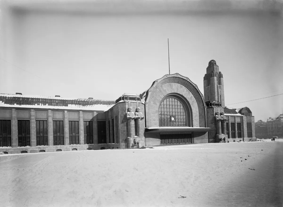 Fotografia em preto e branco da fachada da estação sob neve, com grande arco envidraçado, torre e figuras de pedra diante da entrada.