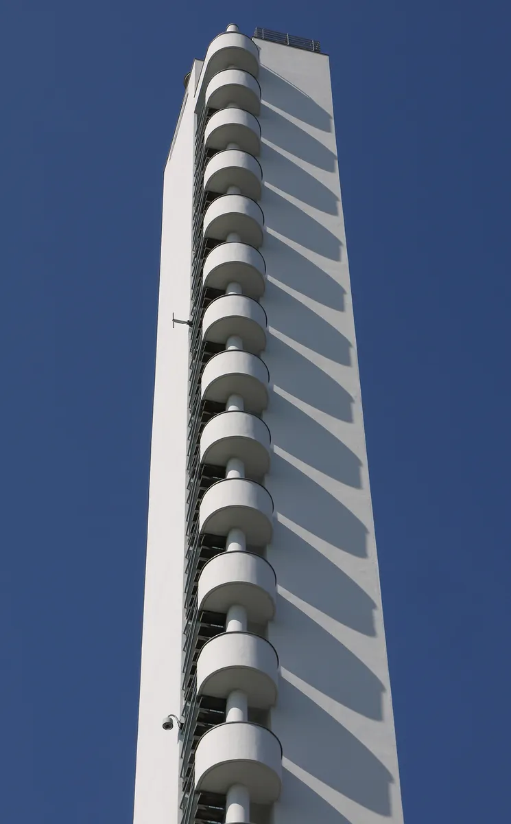 Parte superior da torre branca vista de baixo, com escada externa curva, corrimãos e céu azul.