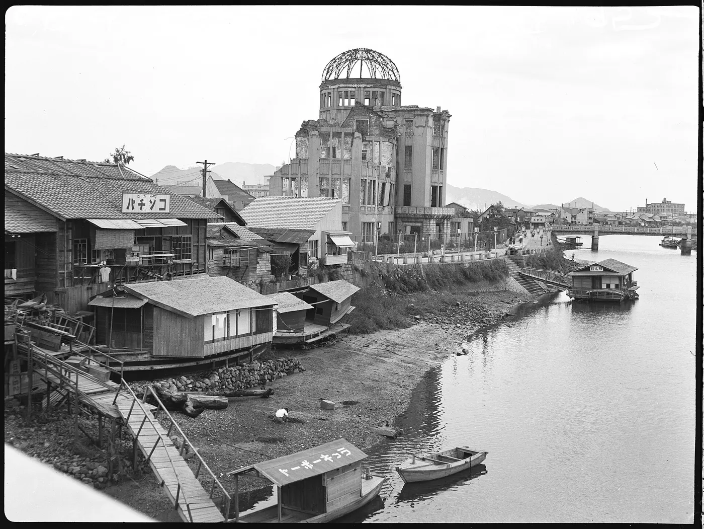 Um prédio arruinado com a cúpula de ferro exposta, junto a casas de madeira na margem de um rio.