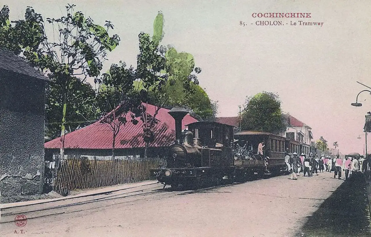 Cartão-postal colorido mostrando uma pequena locomotiva a vapor puxando vagões de um bonde em Chợ Lớn, com pedestres ao lado da via.