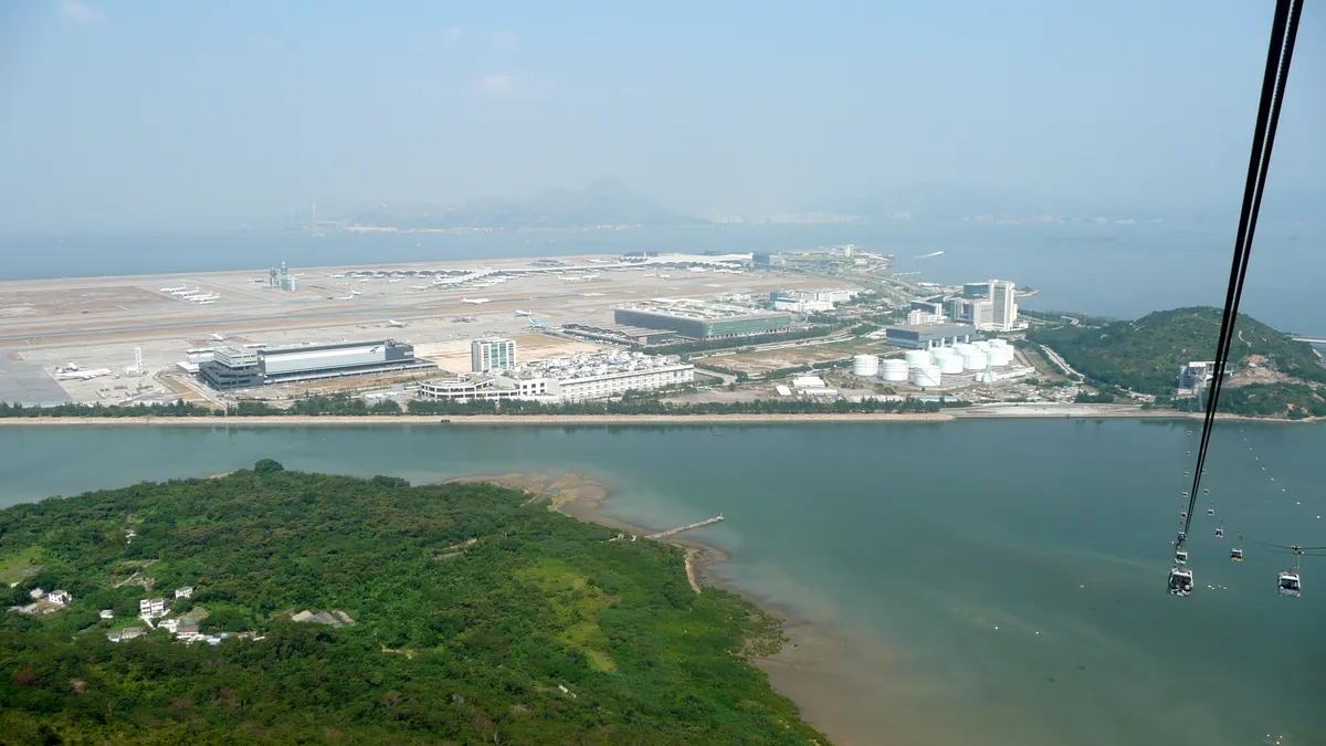 Ilha aeroportuária com pistas, um terminal de arcos e aviões, vista do alto, com o mar e as montanhas ao fundo.