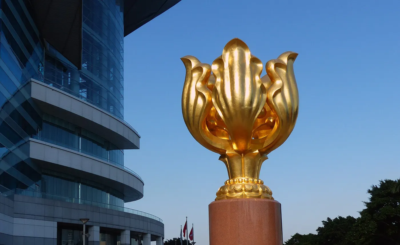 Uma flor dourada de metal sobre um pedestal, numa praça vazia à beira da água.