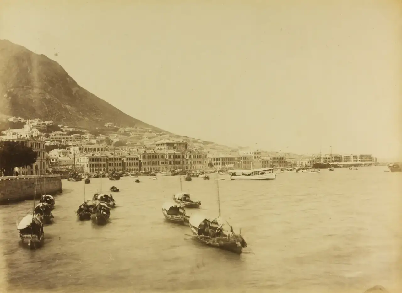 Fotografia em sépia da orla de Hong Kong com fileiras de edifícios coloniais ao pé de uma montanha, e várias juncas ancoradas nas águas em primeiro plano.
