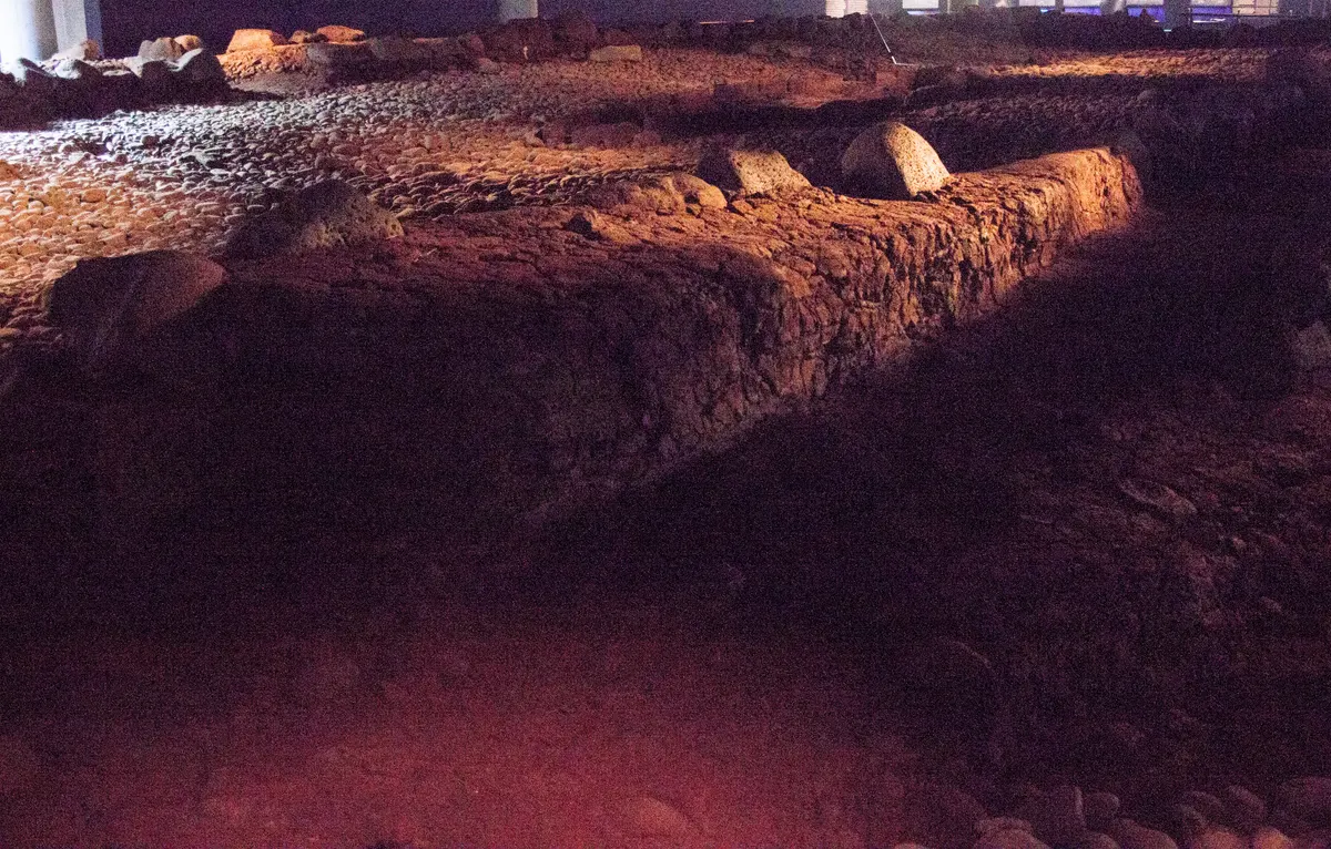 Lareira alongada escavada, com bordas de terra e pedras, no piso escuro da casa preservada no museu.