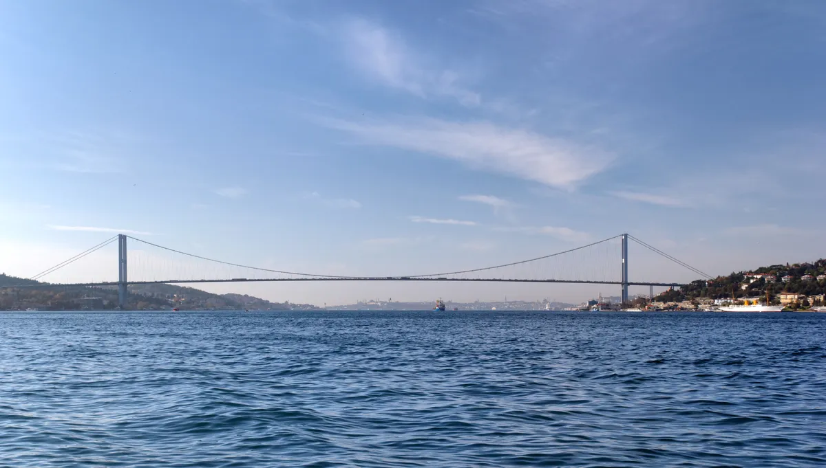 Ponte suspensa com torres e cabos sobre a água, margens urbanizadas, embarcações e céu azul com nuvens.
