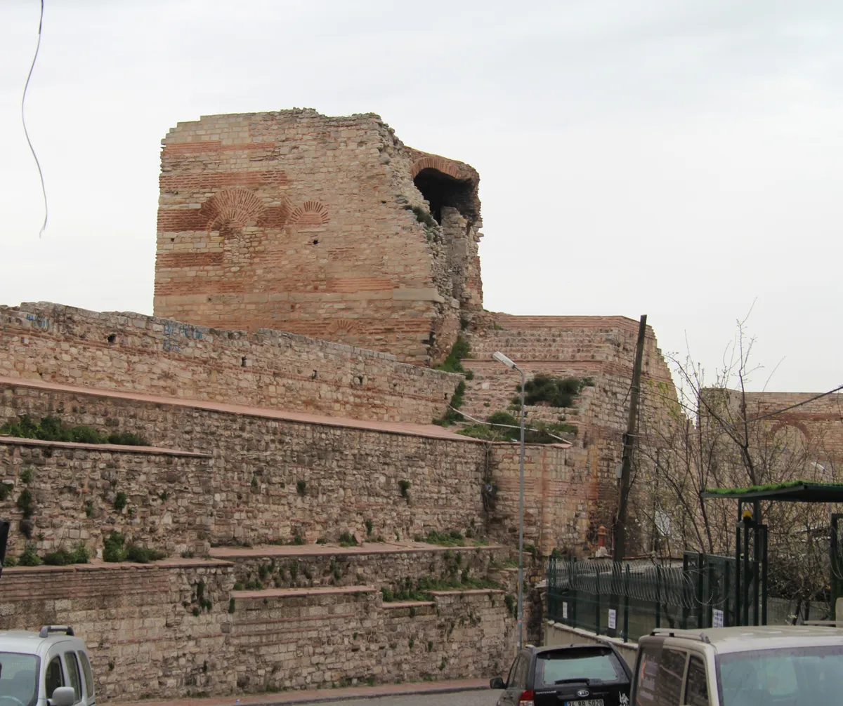 Torre parcialmente arruinada e muros de pedra e tijolo, com vegetação, grades e partes de veículos na faixa inferior.