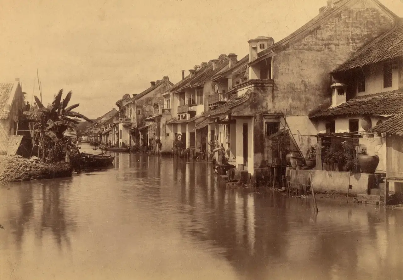 Fotografia em sépia de casas com telhados inclinados ao longo de um canal alagado em Batávia, com barcos e moradores nas varandas.