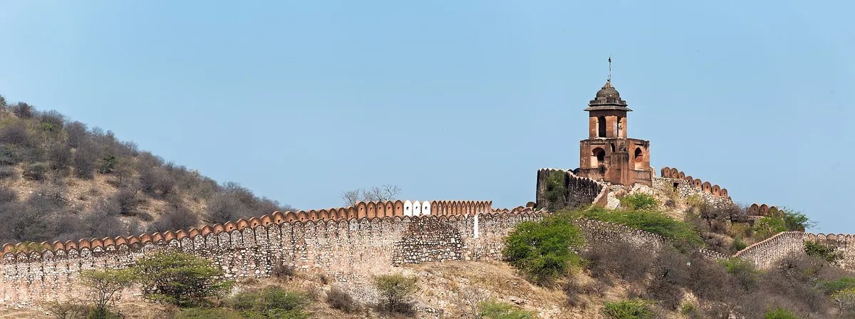 Muralha de pedra com ameias sobre um morro seco, e uma torre de tijolo com cúpula.