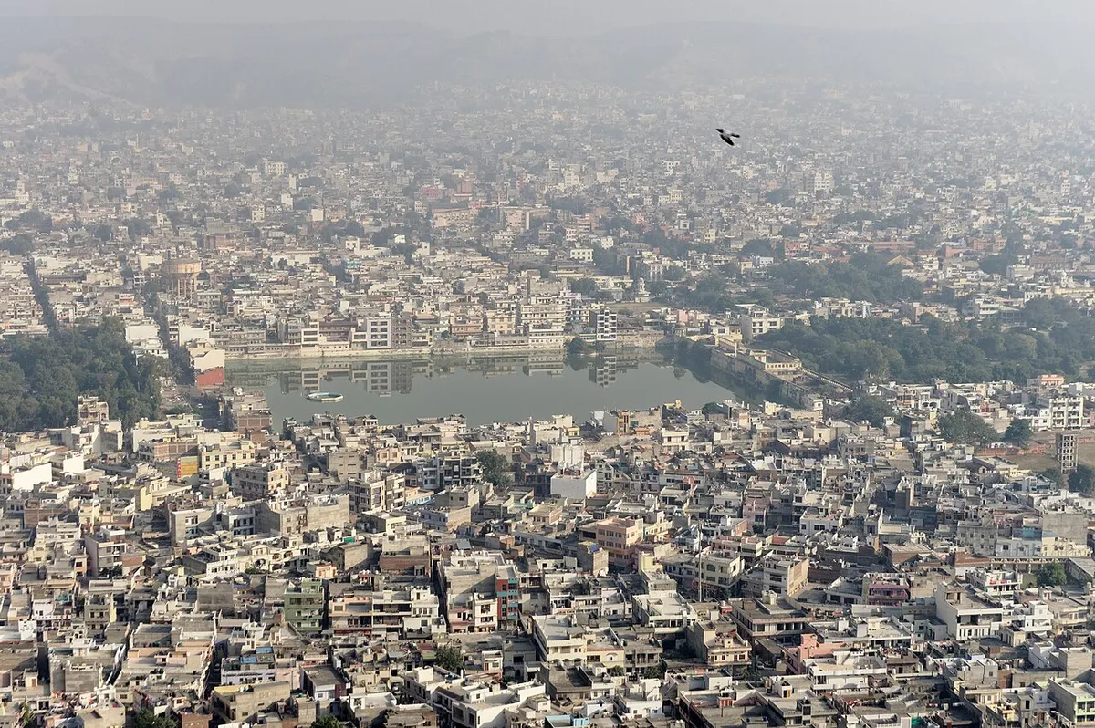 Cidade densa em volta de um lago, com um muro claro na margem e um pássaro no céu.