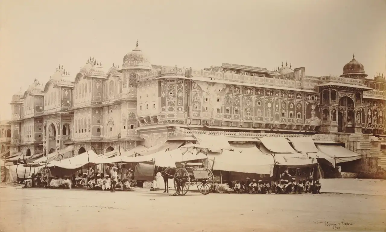 Fotografia em sépia da fachada decorada do Maharaja College em Jaipur, com toldos de mercado, uma carroça puxada por cavalo e pessoas sentadas em frente ao prédio.