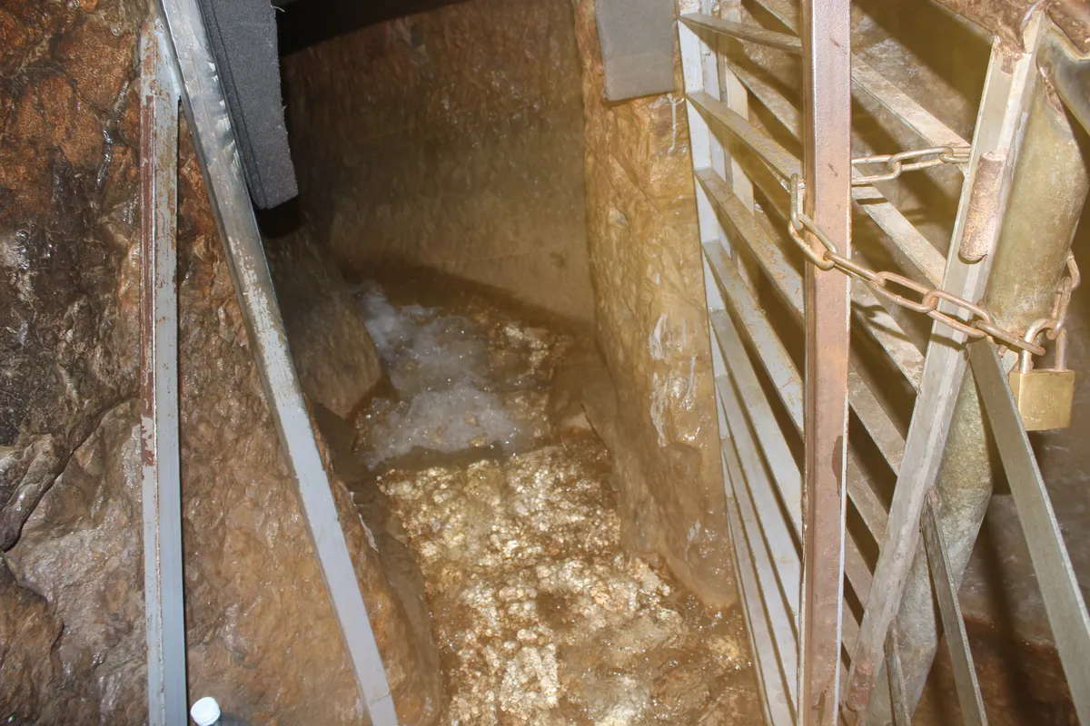Água brota numa abertura rochosa, junto a grades metálicas com corrente e cadeado.