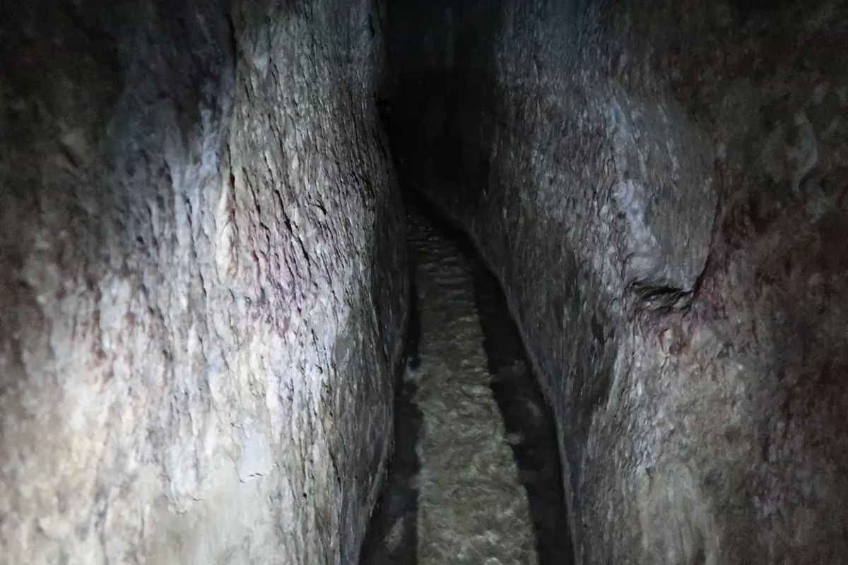 Passagem estreita escavada em rocha, com água no chão e interior escuro ao fundo.