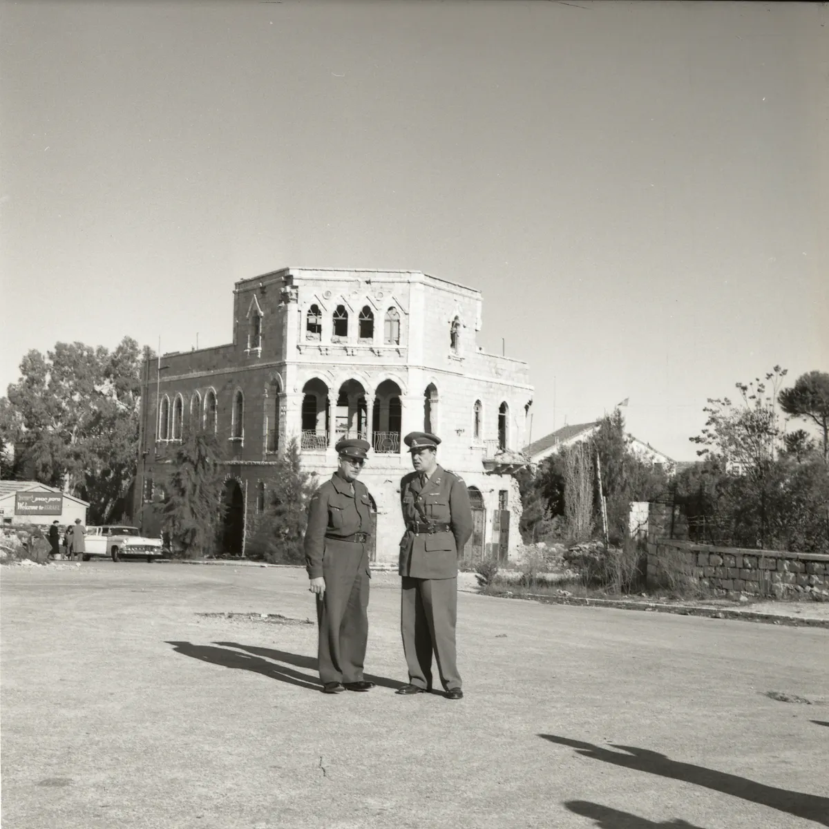 Dois homens de uniforme diante de um edifício de pedra com arcos, numa rua com carro e placa ao fundo.