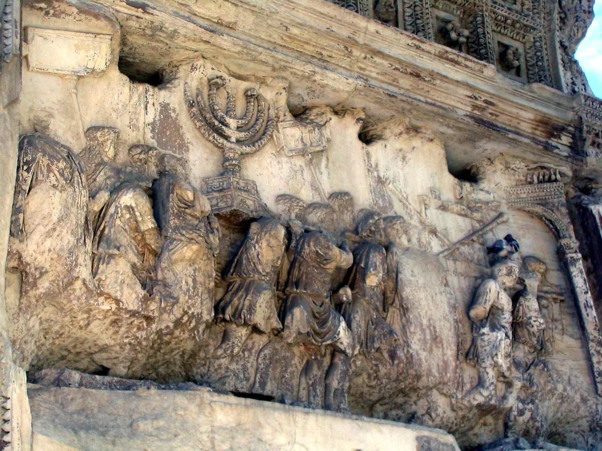 Relevo antigo em pedra com uma procissão que carrega um candelabro, junto a um arco esculpido; figuras desgastadas e frisos nas bordas.