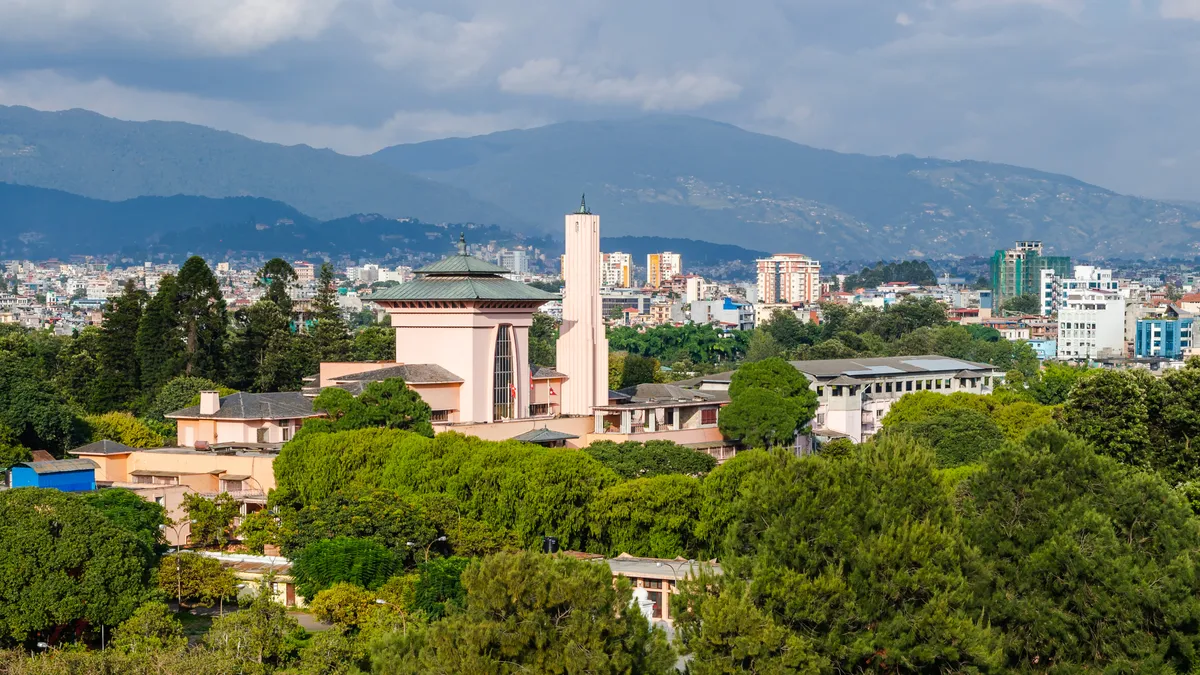 Palácio rosa com telhado verde e uma torre alta, visto por cima das árvores, com a cidade e as montanhas ao fundo.
