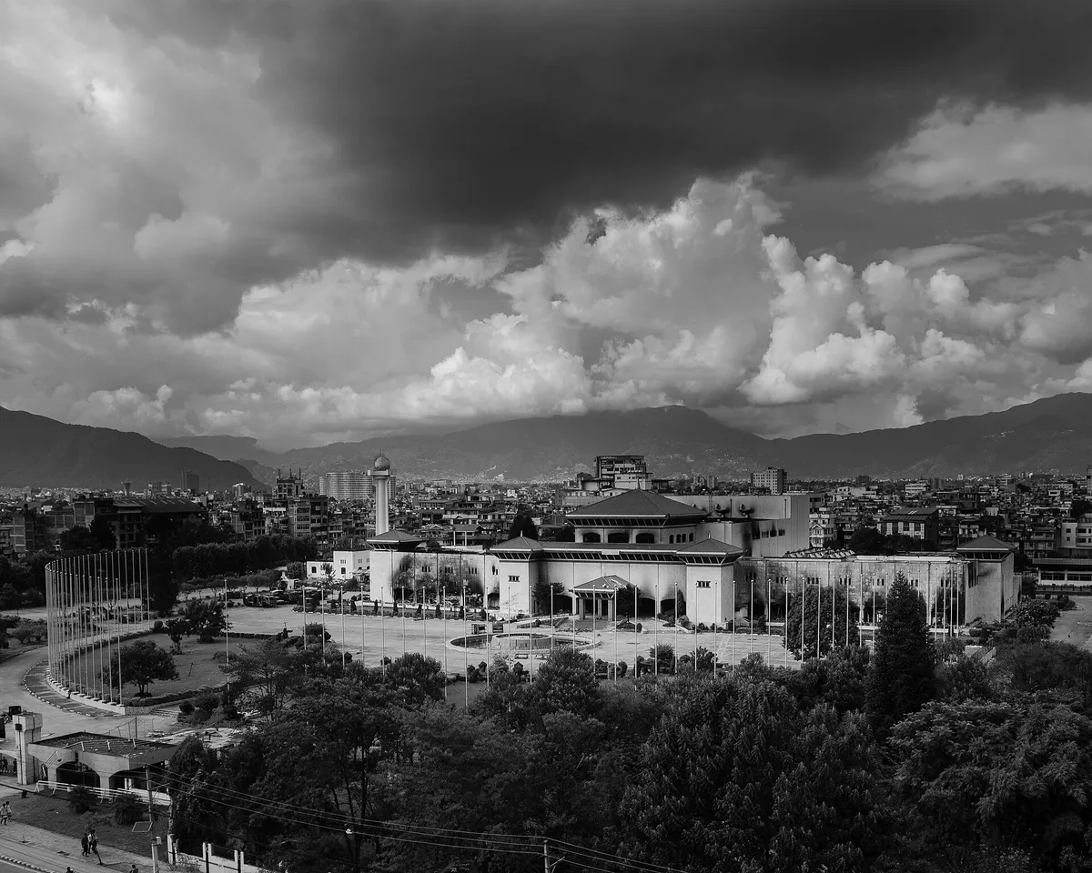 O edifício de telhados em degraus, com manchas escuras no telhado, a praça e as montanhas ao fundo.
