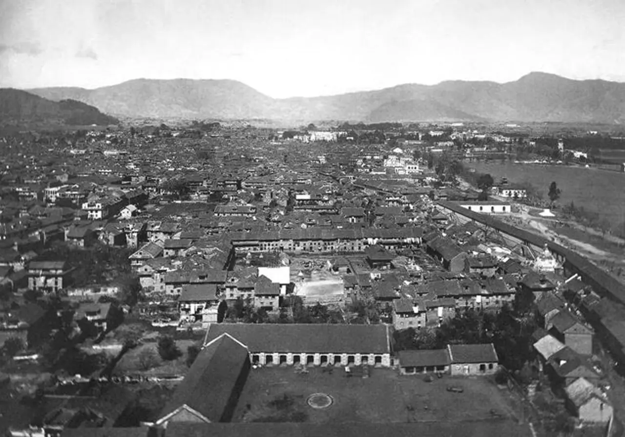 Fotografia aérea em preto e branco da cidade de telhados juntos, com um prédio comprido e um pátio em primeiro plano, um descampado à direita e morros ao fundo.