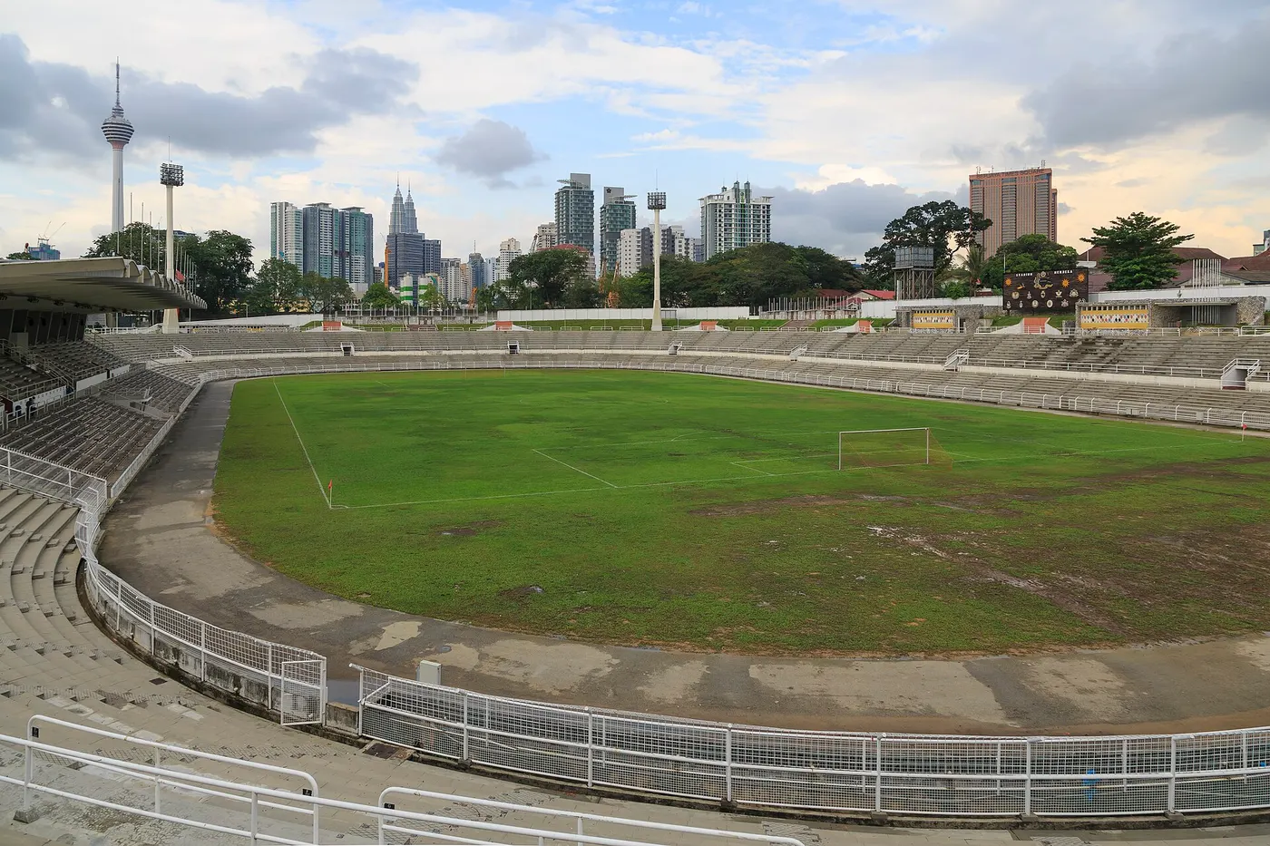 Um estádio oval com gramado, arquibancadas vazias e torres da cidade ao fundo.