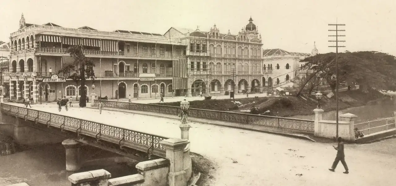 Fotografia em preto e branco de prédios de estilo indo-sarraceno junto a uma ponte, com uma carroça e pedestres atravessando.