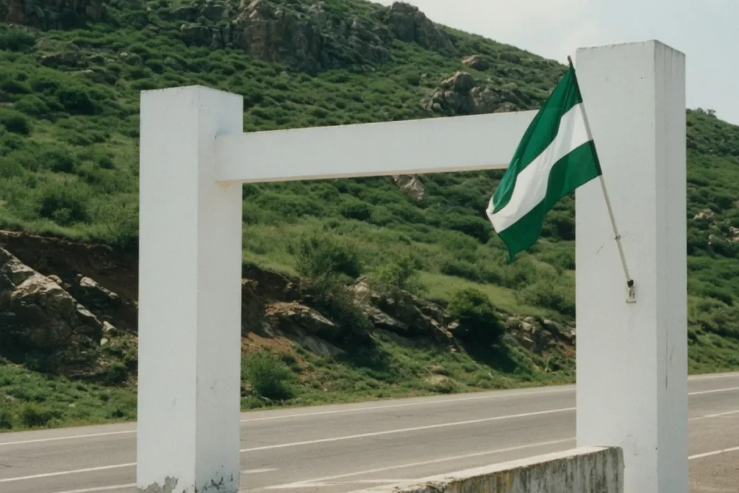 A white gate of two pylons, a green-and-white flag, a rocky hill and an empty road.