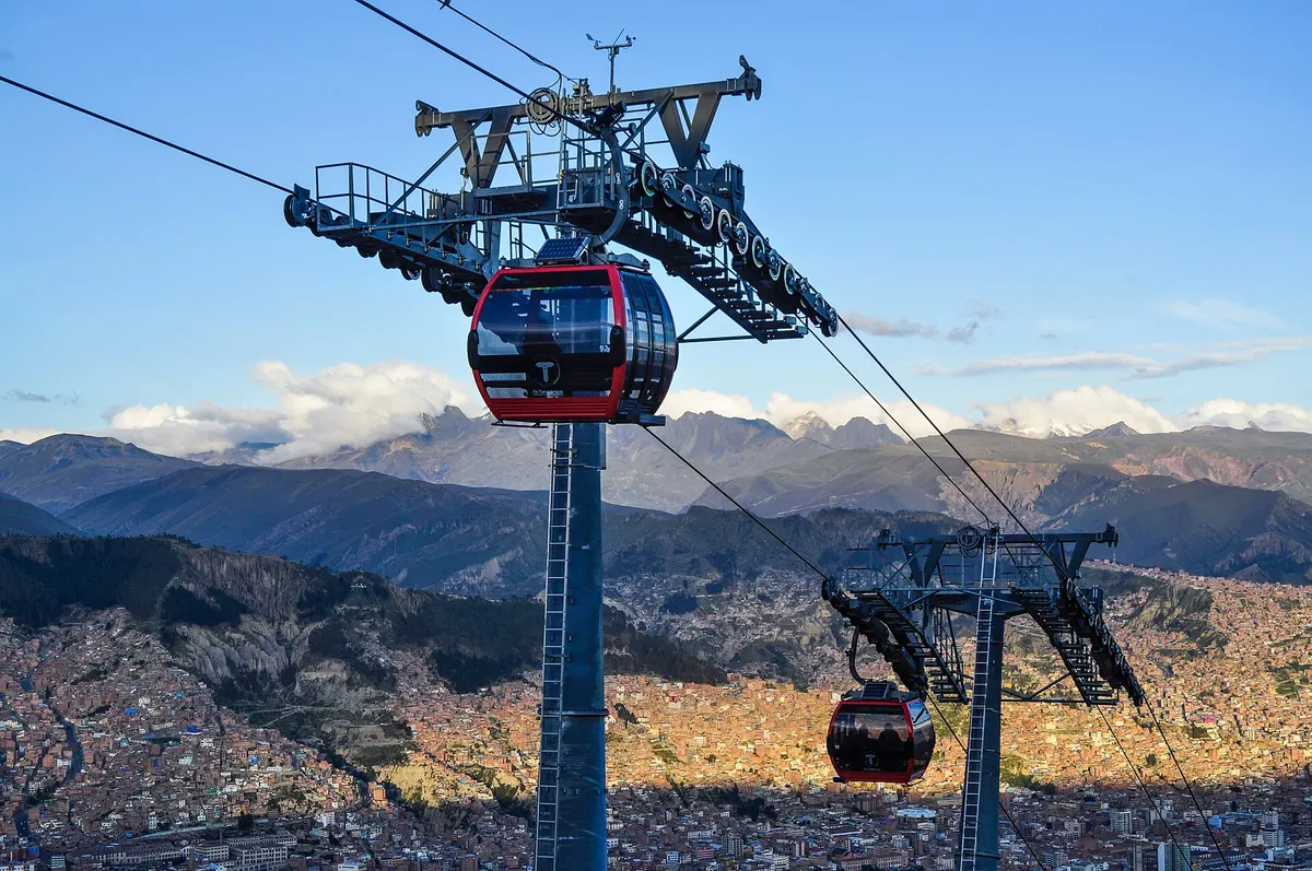 Cabines vermelhas suspensas nos cabos, com a cidade no vale e as montanhas ao fundo.