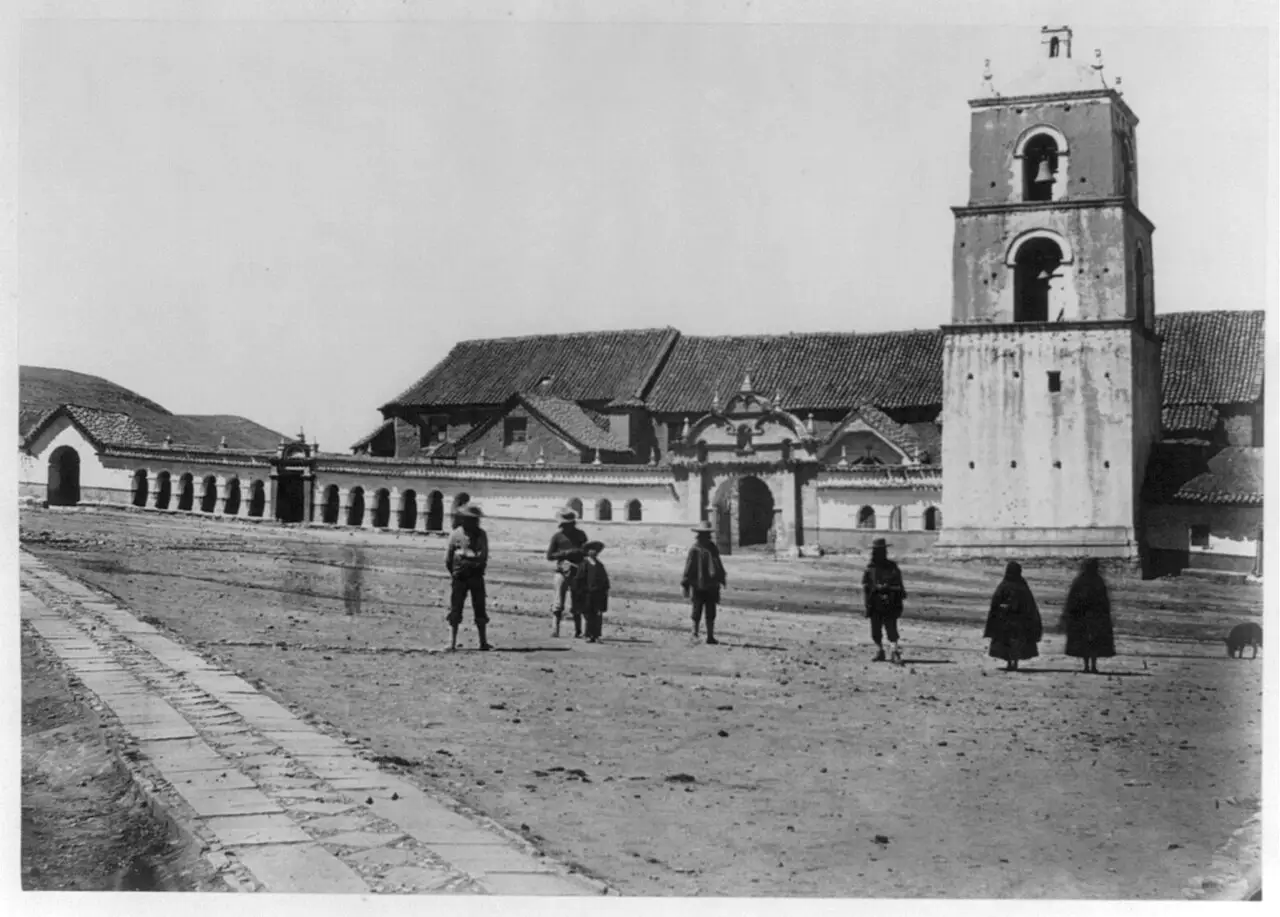 Fotografia em preto e branco da catedral, com a torre à direita, portal e ala de arcadas, e pessoas espalhadas numa praça de terra.