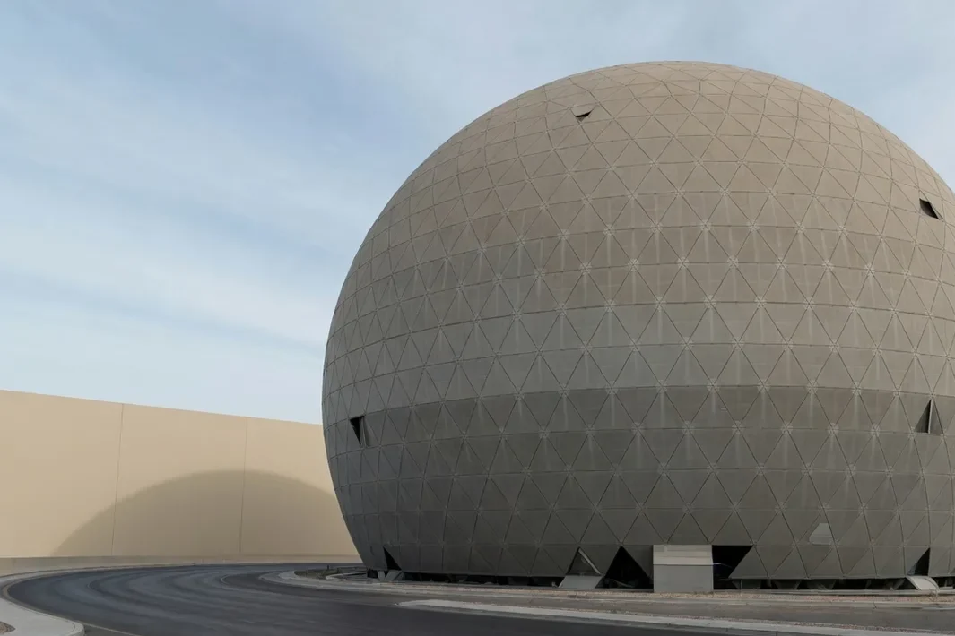 A esfera bege de painéis triangulares à direita, com o céu azul claro no meio e a rua vazia.