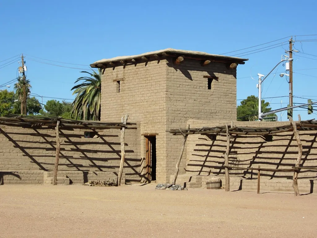 Uma torre e um muro de adobe, com um telheiro de troncos na frente.