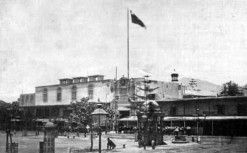 Um palácio baixo numa praça, com uma bandeira num mastro alto e figuras pequenas demais para reconhecer.