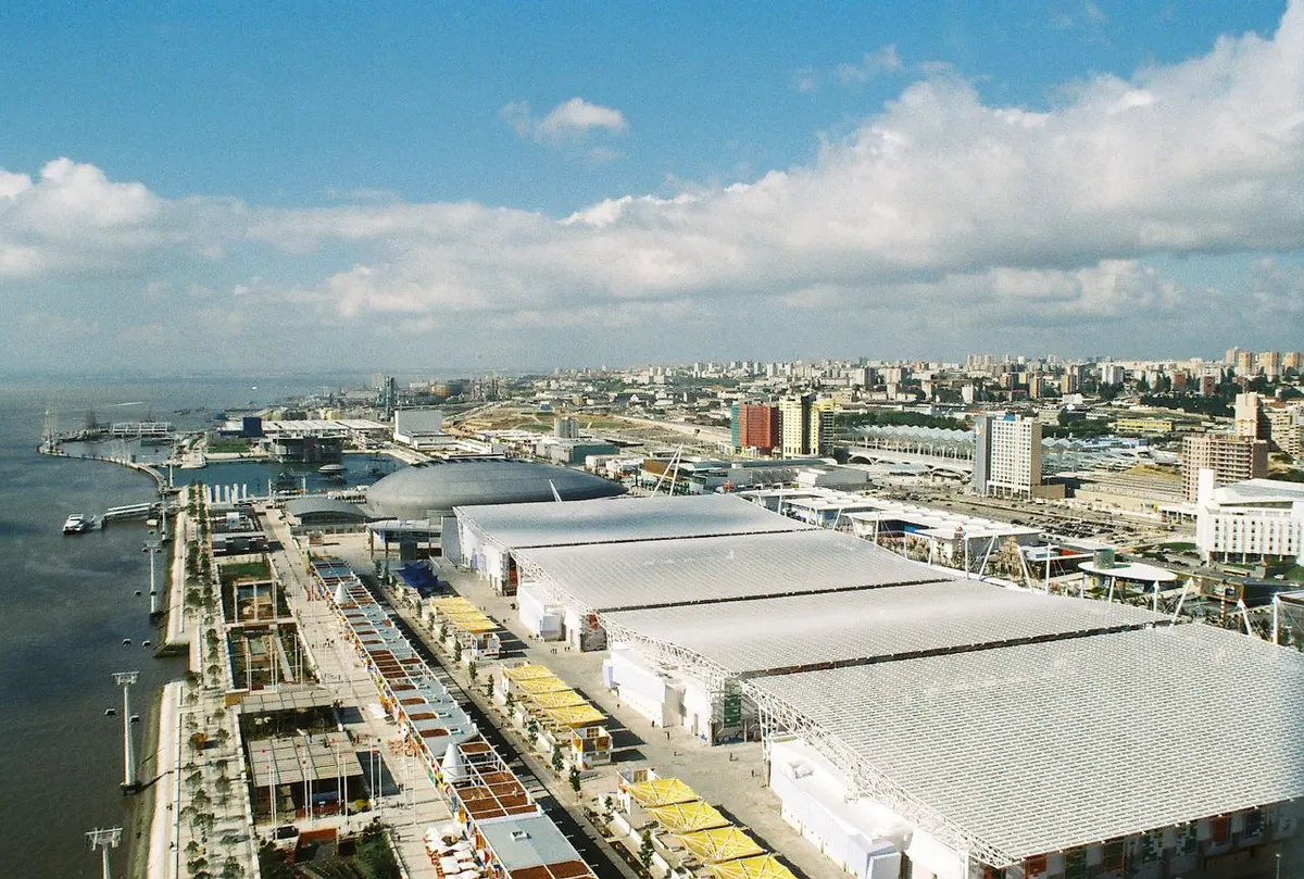 Vista elevada da Expo ’98 junto ao Tejo, com pavilhões de grandes coberturas claras, teleférico, caminhos e edifícios do recinto.
