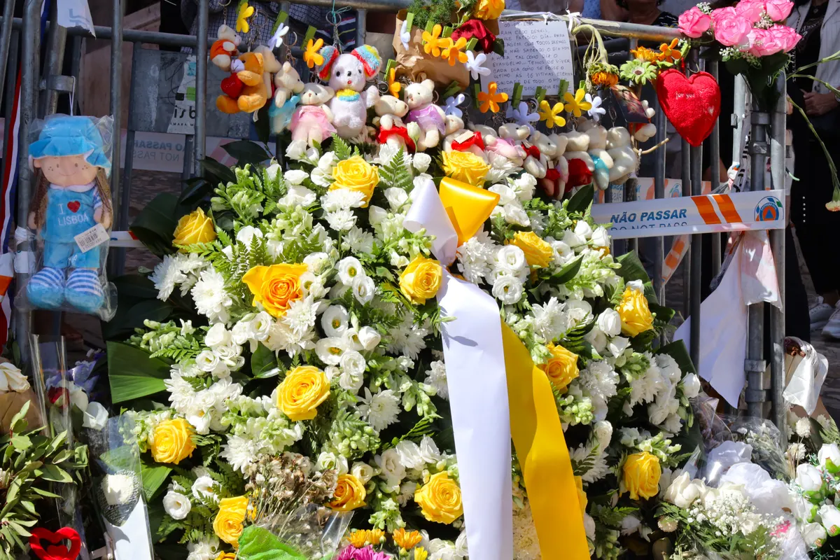 Flores brancas e amarelas, fitas, mensagens e bonecos junto às grades e à fita de isolamento no memorial às vítimas do acidente da Glória.