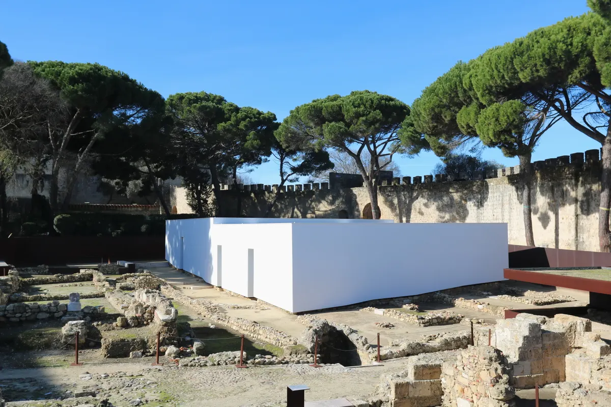 Núcleo arqueológico no Castelo de São Jorge, com muros de pedra, caminhos de visita e o pavilhão branco que protege as casas islâmicas, sob árvores e muralha.