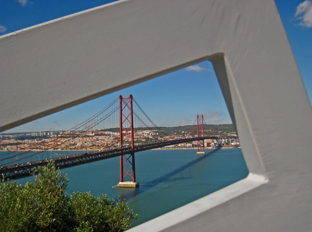Ponte suspensa vermelha sobre o Tejo e Lisboa ao fundo, vista através de uma abertura numa estrutura branca na margem de Almada.