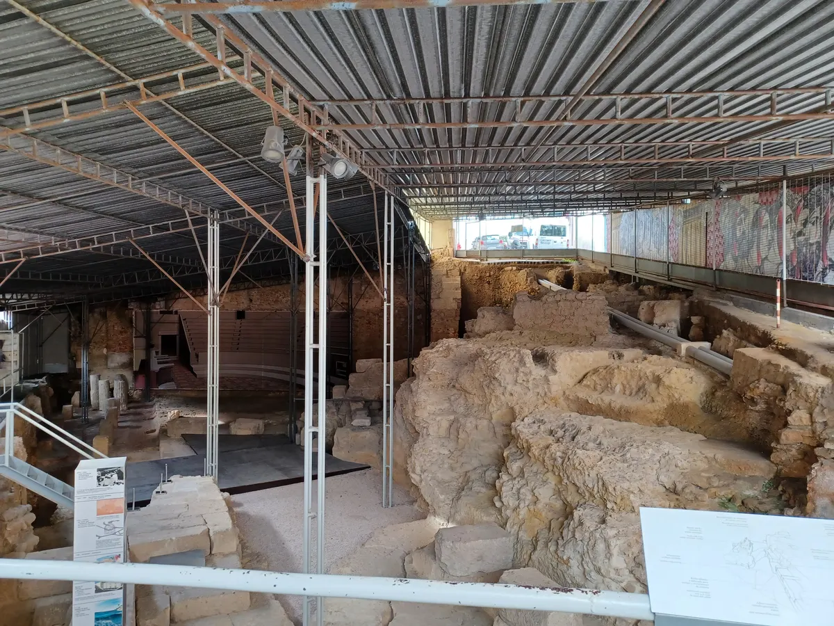 Ruínas do teatro romano sob cobertura metálica, com degraus, muros escavados, passarelas e painéis de visitação.