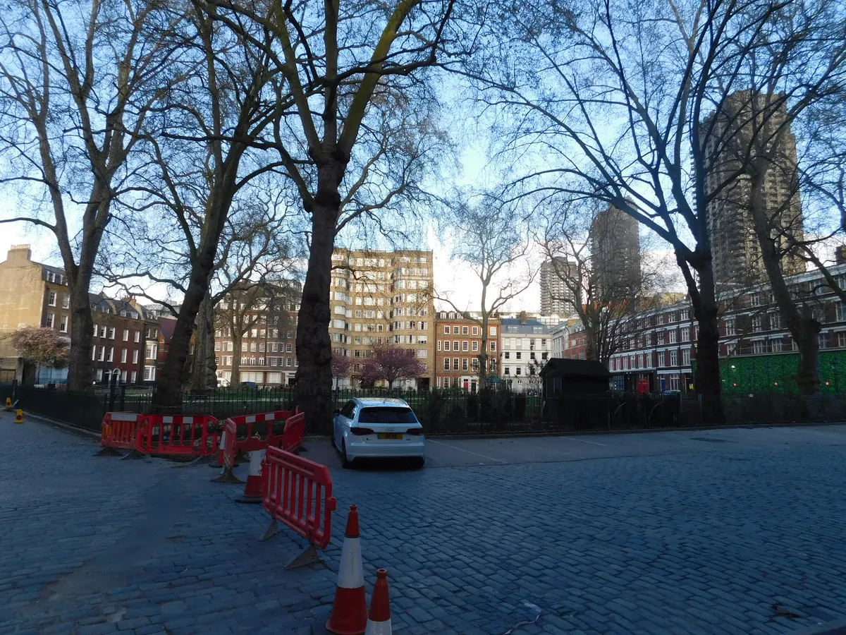 Jardim cercado de Charterhouse Square, com árvores sem folhas, prédios, barreiras vermelhas e um carro ao fundo.