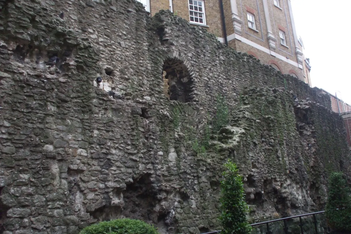 Trecho de muralha de pedra e tijolos, com vegetação, pombos e edifícios ao fundo.