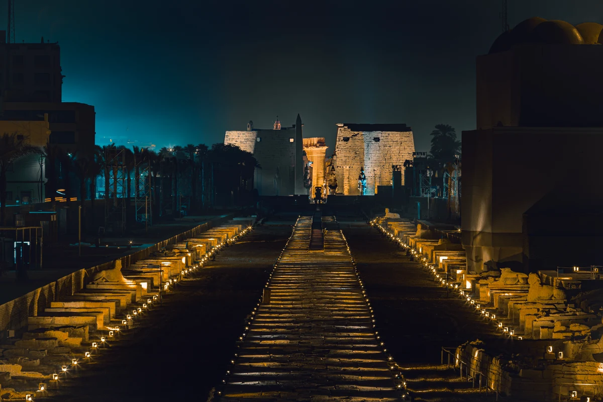 Caminho iluminado entre fileiras de esfinges, com os pilonos do templo ao fundo e edifícios nas laterais.