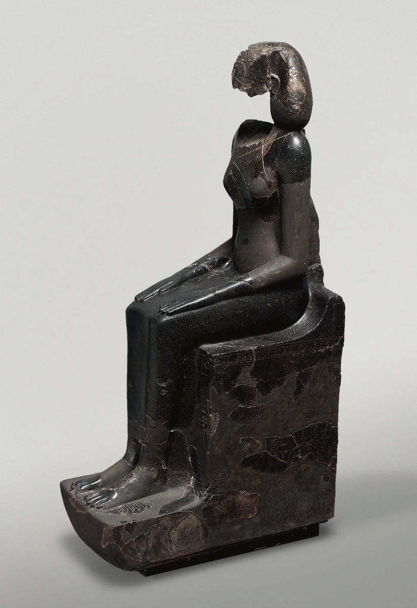 Estátua escura sentada, com rosto fragmentado, mãos sobre os joelhos e trono com inscrições.