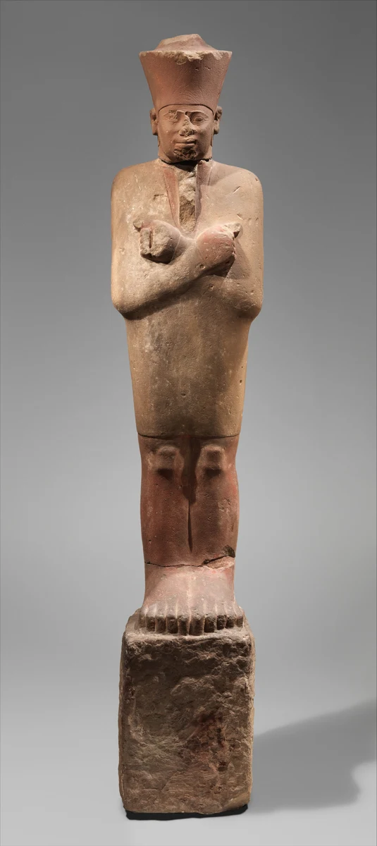 Estátua de pedra avermelhada com coroa danificada, braços cruzados, pés e base áspera visíveis.